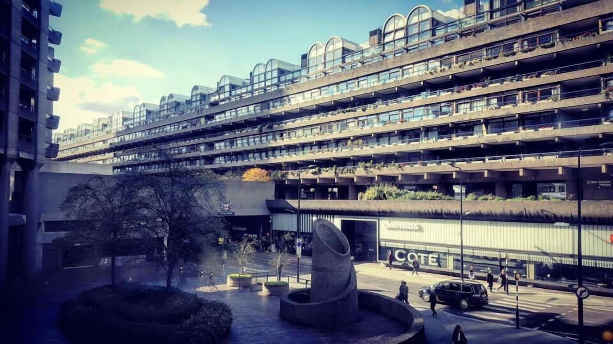 Montcalm Brewery Townhouse Hotel London Exterior photo The photo shows a modern architectural structure with multiple levels, featuring a series of large, rectangular balconies. Below this building, there is a plaza or courtyard area with a sculptural piece, which appears to be made of stone. Surrounding