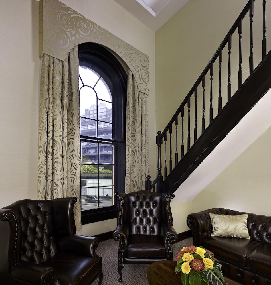 Montcalm Brewery Townhouse Hotel London Exterior photo The photo shows a cozy interior space featuring two elegant leather armchairs placed near a large, ornate window with arched top and decorative curtains. The wall is painted in a muted color, enhancing the warmth of the room. A staircase with black s