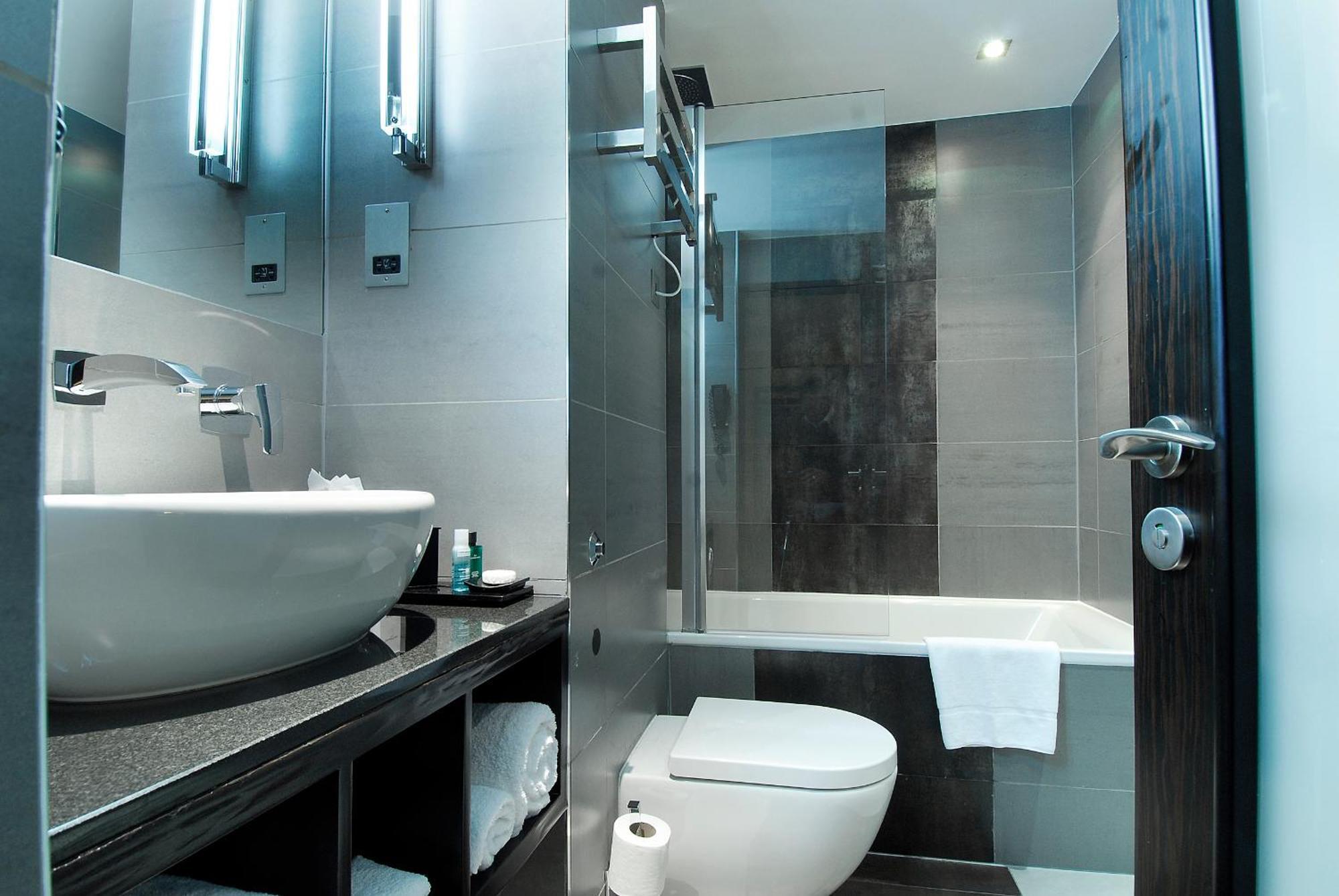 Montcalm Brewery Townhouse Hotel London Exterior photo The photo shows a modern bathroom with sleek design elements. It features a curved white sink on a dark countertop, with a mirror above that is illuminated by stylish lighting fixtures. Next to the sink, there's a toilet, and on the opposite side, a 
