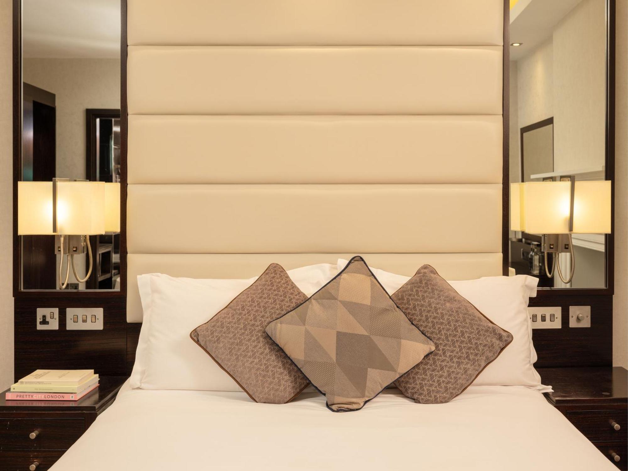 Montcalm Brewery Townhouse Hotel London Exterior photo The image shows a cozy bedroom setting. The bed features a headboard upholstered in a light beige fabric. It is adorned with three decorative pillows in varying patterns and colors, placed on white bedding. On either side of the bed, there are modern