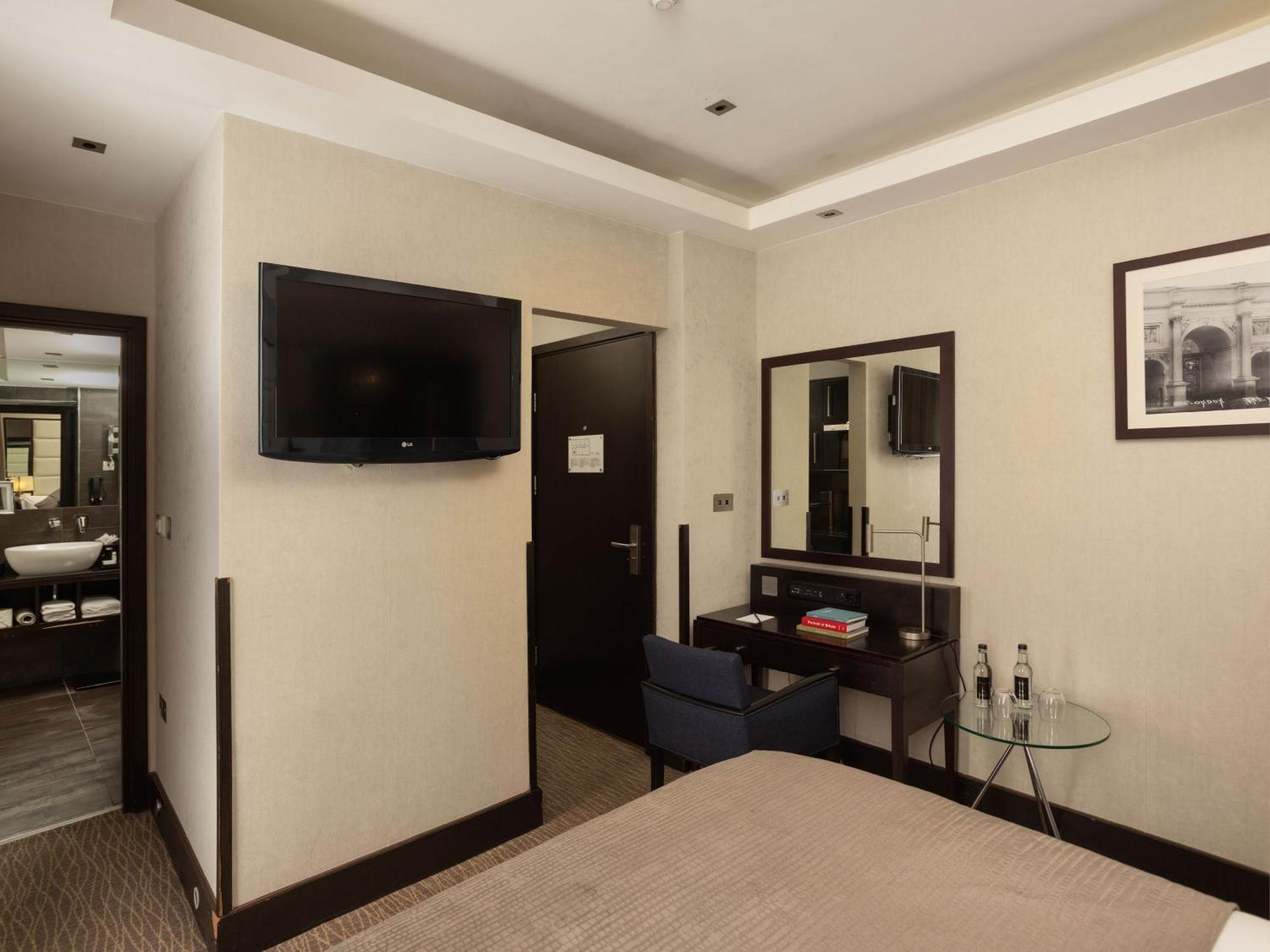 Montcalm Brewery Townhouse Hotel London Exterior photo The photo shows a hotel room with a modern and minimalist design. On the left, there's a wall-mounted flat-screen TV. In the center, there's a bed with a neutral-colored bedspread. To the right of the bed, there is a small desk with a mirror above it