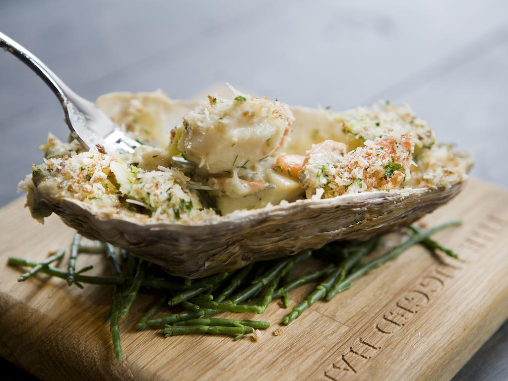 Montcalm Brewery Townhouse Hotel London Exterior photo The photo shows a large oyster shell filled with a mixture that appears to include seafood, herbs, and breadcrumbs. The dish is presented on a wooden board with some greenery, possibly a type of seaweed or garnishing. A fork is placed beside the oyst