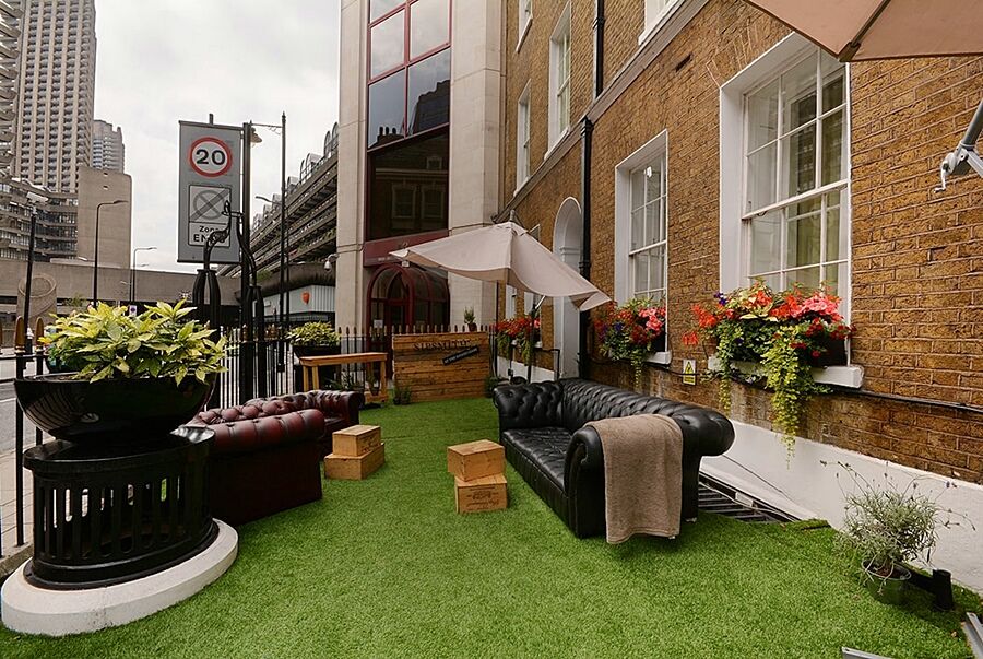 Montcalm Brewery Townhouse Hotel London Exterior photo The photo depicts a charming outdoor space, likely a patio or garden area. It features two comfortable-looking leather sofas, one of which is a deep burgundy color, placed on a green artificial lawn. There are wooden crates that could serve as side t